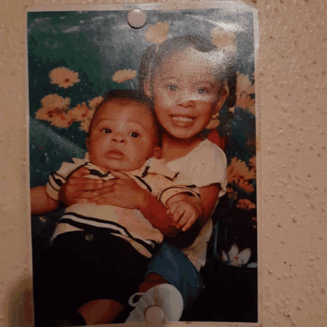 a picture of a little girl holding a baby with flowers in the background