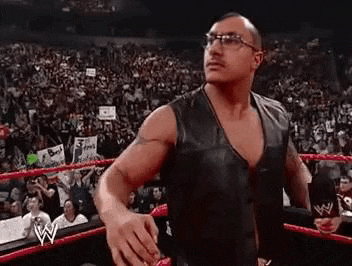 a man wearing glasses and a leather vest is standing in a wrestling ring in front of a crowd .