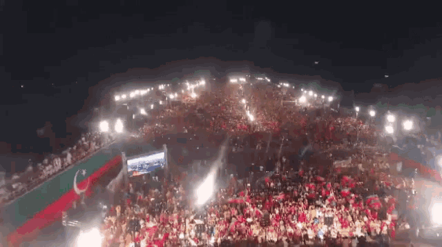 a large crowd of people are gathered in a stadium at night with a green and red flag behind them