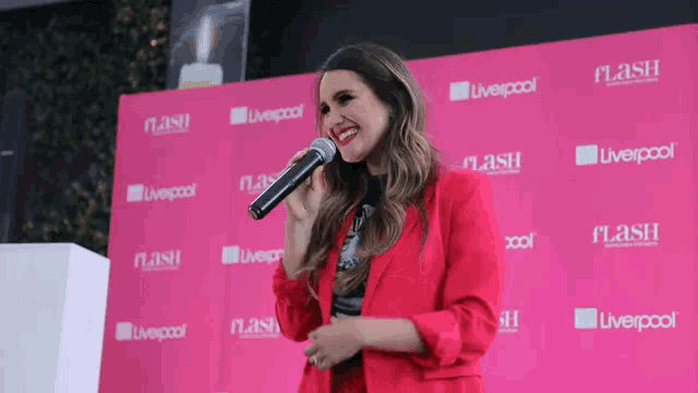 a woman in a red jacket sings into a microphone in front of a pink wall that says liverpool