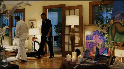 two men are standing in a living room with candles and a painting on the table