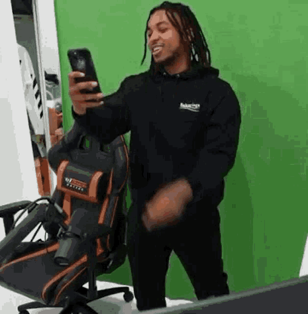 a man is standing in front of a green screen holding a cell phone and a chair .