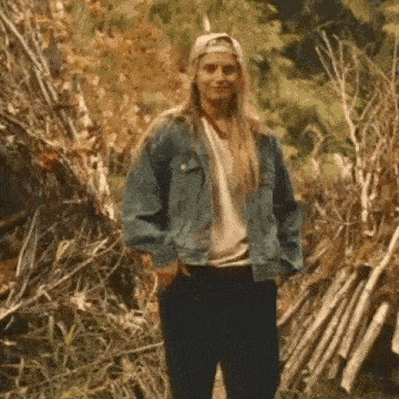 a woman wearing a denim jacket and a baseball cap is standing in front of a pile of branches .