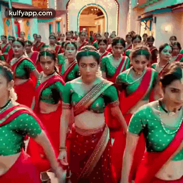 a group of women in red and green saris are dancing in a line .