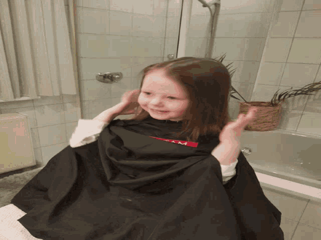 a little girl is getting her hair cut in a bathroom with a black cape on
