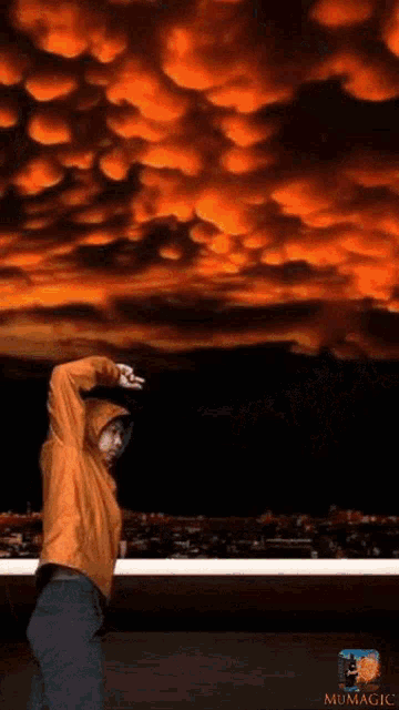 a man in an orange hoodie stands in front of a cloudy sky with mumagic written on the bottom right