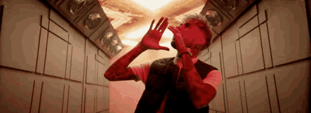 a man in a red vest is standing in a dark room with his hands on his face