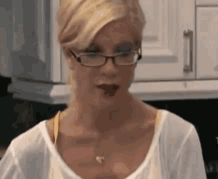 a woman wearing glasses is standing in a kitchen with a glass of wine in her hand .