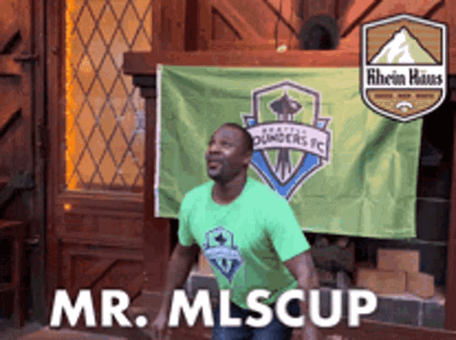 a man in a green shirt is standing in front of a mr. mlscup sign