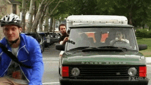 a man wearing a helmet is riding a bike next to a green range rover