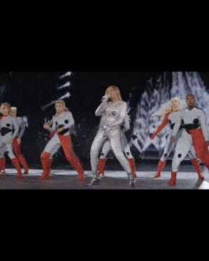 a woman in a silver bodysuit is surrounded by a group of dancers