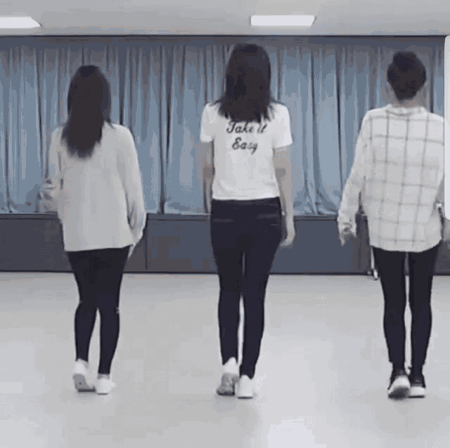 three women are walking in a room with one wearing a t-shirt that says take it easy on the back .