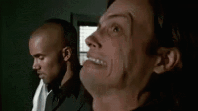 two men are standing next to each other in a room . one of the men is making a funny face .