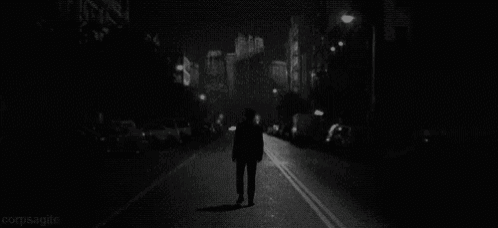 a black and white photo of a man walking down a city street at night .