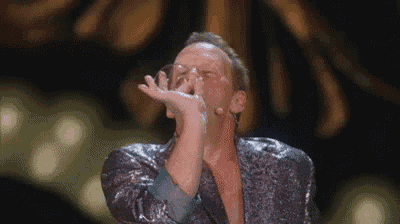 a man in a sequined jacket is singing into a microphone while holding his hand over his mouth .