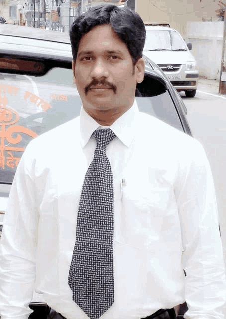 a man in a white shirt and tie is standing in front of a car with a license plate that says rj 5852