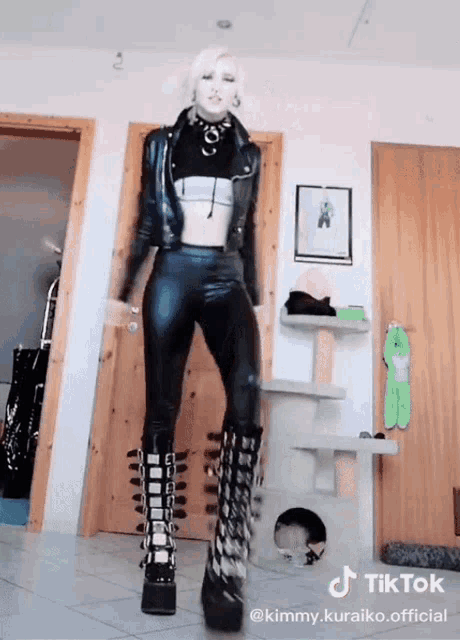 a woman wearing a leather jacket and pants is dancing in a room with a cat tree behind her