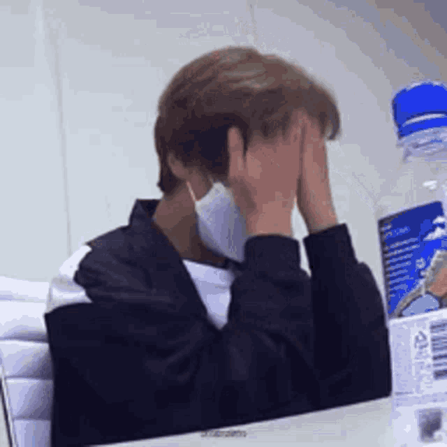 a man wearing a mask is sitting at a desk with his hands on his face .