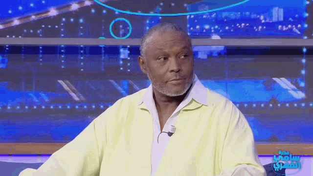a man in a yellow shirt is sitting in front of a screen that says ' arabic sports ' on it