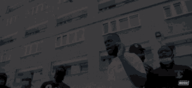 a group of men are standing in front of a building and one of them is wearing a mask with the letter m on the back of his shirt