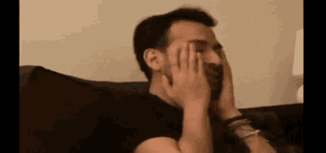 a man is sitting on a couch and covering his face with his hands .
