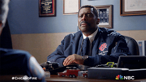 a man in a chicago fire jacket sits at a desk in front of a nbc logo