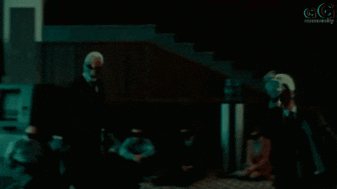 a man in a suit and tie is standing in a dark room with the letters gg in the corner