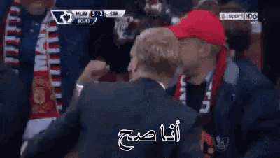 a man in a suit stands in a crowd watching a soccer game with arabic writing on the screen