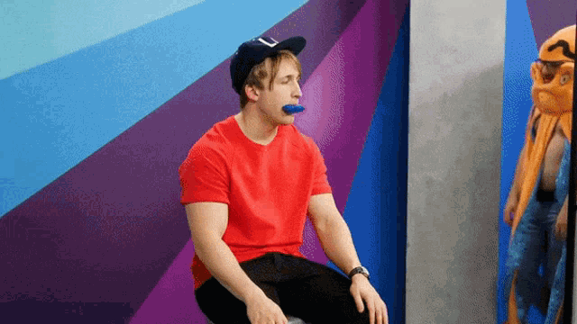 a man wearing a red shirt and a black hat with the letter j on it is sitting in front of a colorful wall