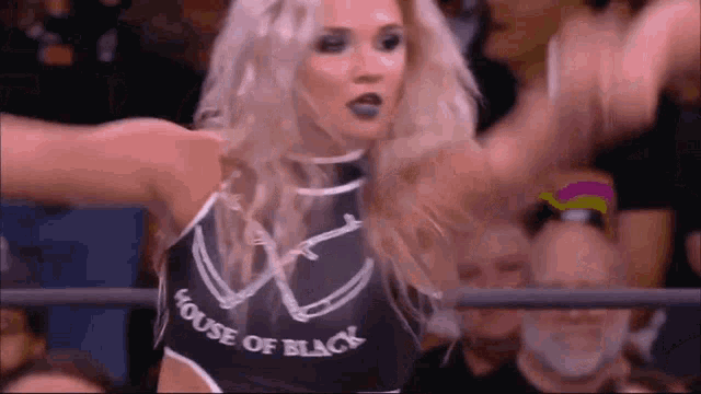 a woman in a black tank top with the word house of black on it is standing in a ring with her arms outstretched .