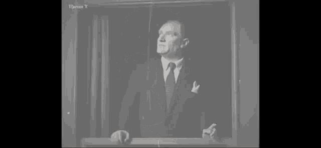 a black and white photo of a man in a suit and tie standing in front of a window .