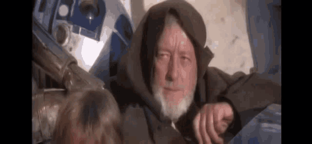 a man with a beard and a hooded jacket is sitting next to a robot .