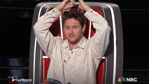 a man in a white shirt is sitting in a chair with his hands in his hair and a nbc logo behind him