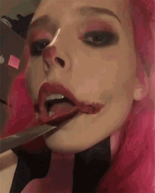 a woman with pink hair is holding a knife in her mouth and making a joker face .