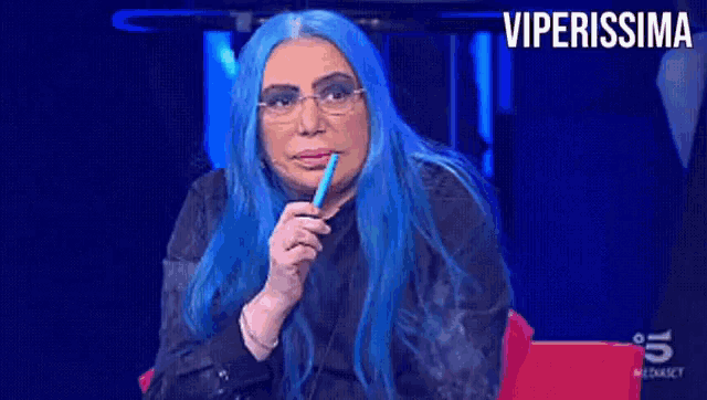 a woman with blue hair and glasses is smoking a cigarette in front of a screen that says viperissima on it