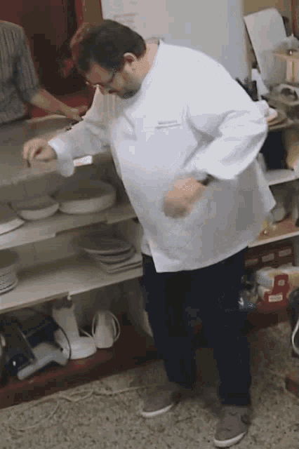 a man in a chef 's coat is standing in front of a shelf with plates on it