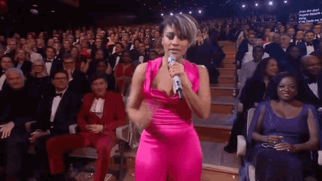 a woman in a pink jumpsuit is singing into a microphone in front of a crowd of people