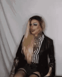a woman is sitting in a chair with her legs crossed and wearing a black jacket and striped shirt .