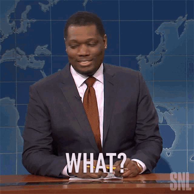 a man in a suit and tie is sitting at a desk with a pen in his hand and says what