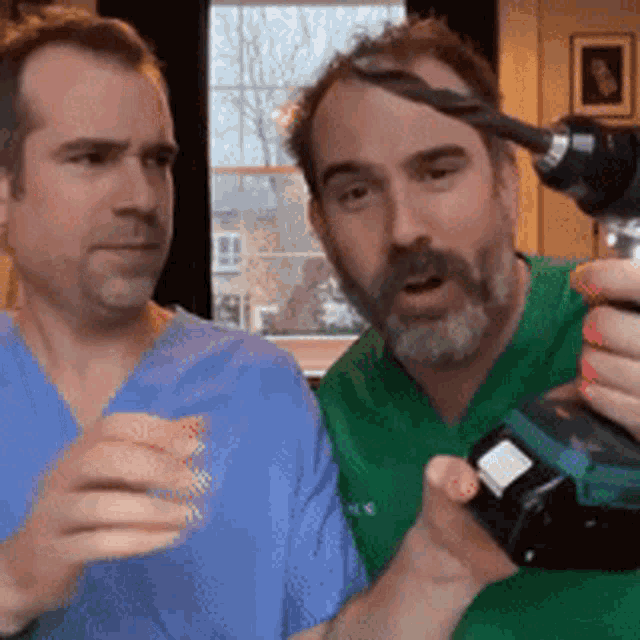 a man in a green shirt is holding a drill next to another man in a blue scrub