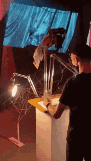 a man in a black shirt is writing on a piece of paper with a lamp behind him