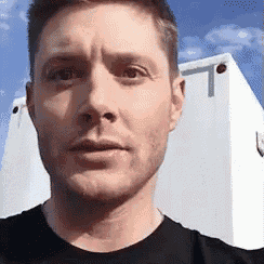 a man in a black shirt is standing in front of a white building and looking at the camera .