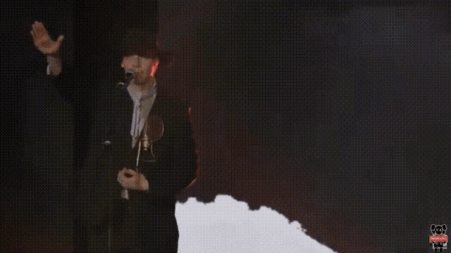 a man in a black suit is singing into a microphone while wearing a hat .