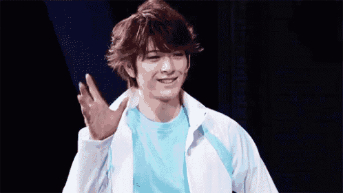 a young man in a white jacket and a blue shirt is waving his hand on a stage .