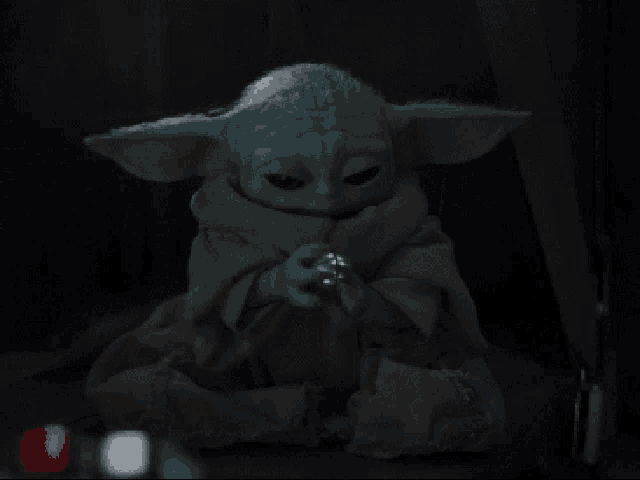 a baby yoda is sitting in a dark room holding a silver object in his hand .