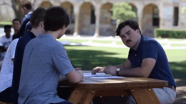 a man with a mustache sits at a picnic table with a group of young men