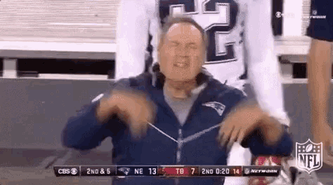 a man in a patriots jacket is sitting in a stadium watching a football game .