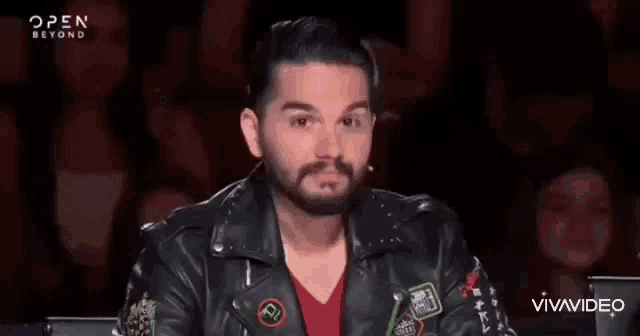 a man with a beard is wearing a leather jacket and making a funny face in front of a crowd .