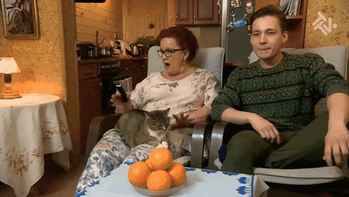 a woman sitting in a chair with a cat and a man sitting in a chair with a bowl of oranges on the table