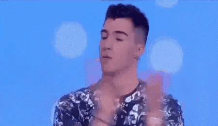 a man in a floral shirt is standing in front of a blue background and talking into a microphone .
